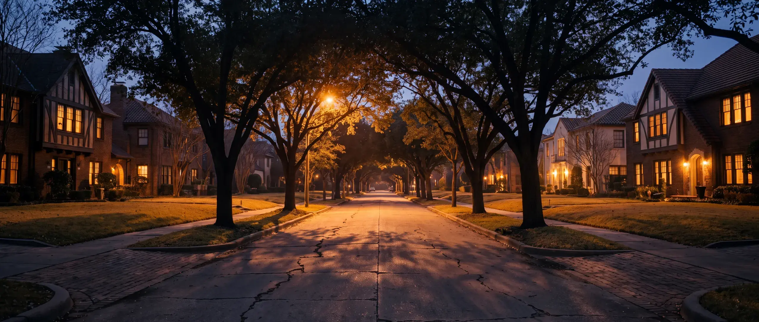 The same East Dallas street at dusk, warm light glowing from the windows of every house.
